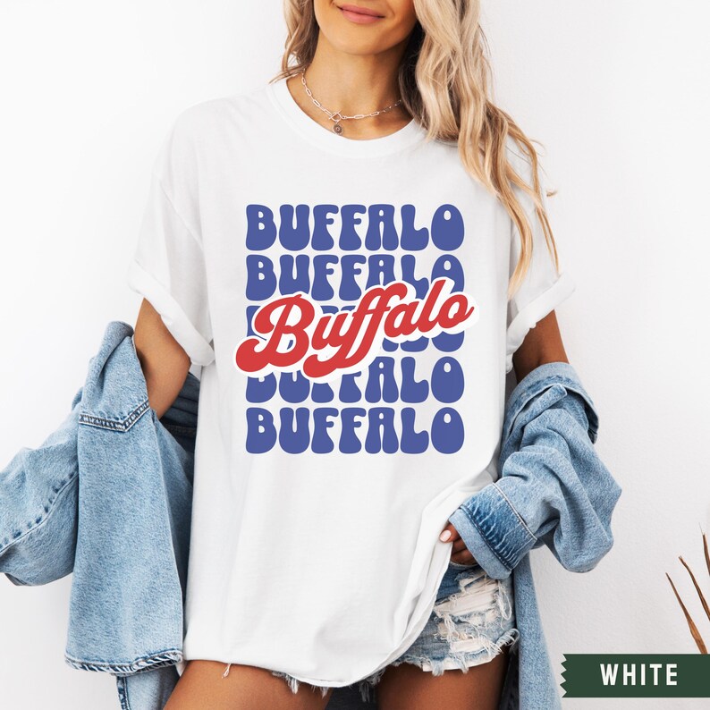 May include: White t-shirt with a retro-style graphic design featuring the word "Buffalo" repeated in blue and red. The word "Buffalo" is in a larger font and red color.