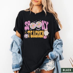 Peut inclure: T-shirt noir avec un motif rose et jaune qui dit "Spooky Teacher" avec un ballon disco et un fant&ocirc;me.