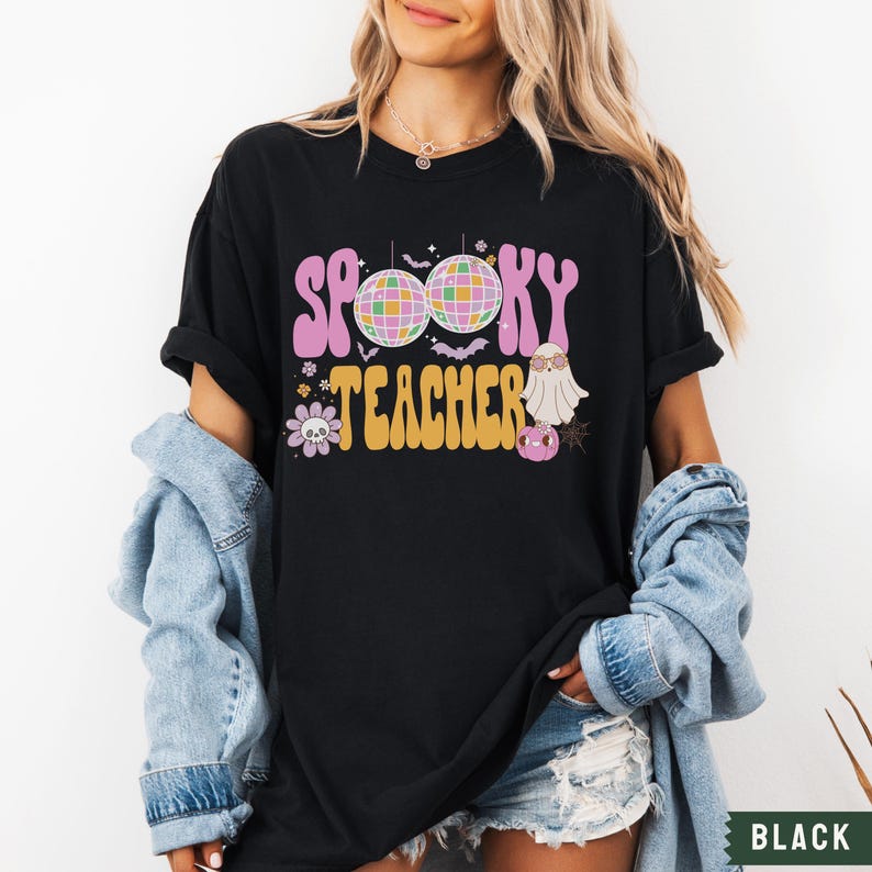 Peut inclure: T-shirt noir avec un motif rose et jaune qui dit "Spooky Teacher" avec un ballon disco et un fant&ocirc;me.