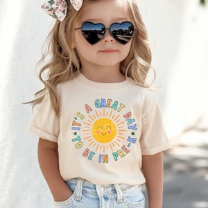 Peut inclure: Un t-shirt &agrave; manches courtes de couleur cr&egrave;me avec un motif de soleil jaune souriant entour&eacute; du texte 'IT'S A GREAT DAY TO BE IN PRE-K' en lettres color&eacute;es et arrondies. Le motif est centr&eacute; sur le devant du t-shirt.