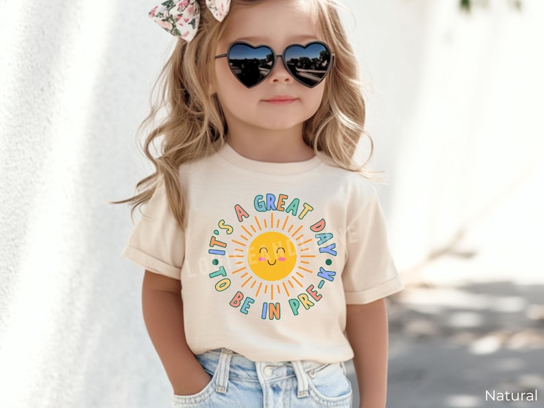 Peut inclure: Un t-shirt &agrave; manches courtes de couleur cr&egrave;me avec un motif de soleil jaune souriant entour&eacute; du texte 'IT'S A GREAT DAY TO BE IN PRE-K' en lettres color&eacute;es et arrondies. Le motif est centr&eacute; sur le devant du t-shirt.
