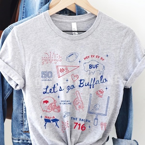 May include: Athletic heather gray t-shirt with a blue and red graphic design. The design includes the text "Let's go Buffalo", a football, a helmet, and other sports-related illustrations. The shirt is hanging on a wooden hanger.