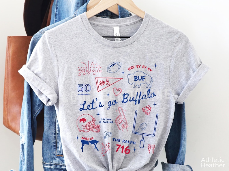 May include: Athletic heather gray t-shirt with a blue and red graphic design. The design includes the text "Let's go Buffalo", a football, a helmet, and other sports-related illustrations. The shirt is hanging on a wooden hanger.