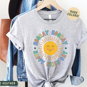 K&ouml;nnte beinhalten: Graues T-Shirt mit einem bunten Sonnen-Motiv und dem Text "Hooray Happy First Day" in einem Kreis um die Sonne.