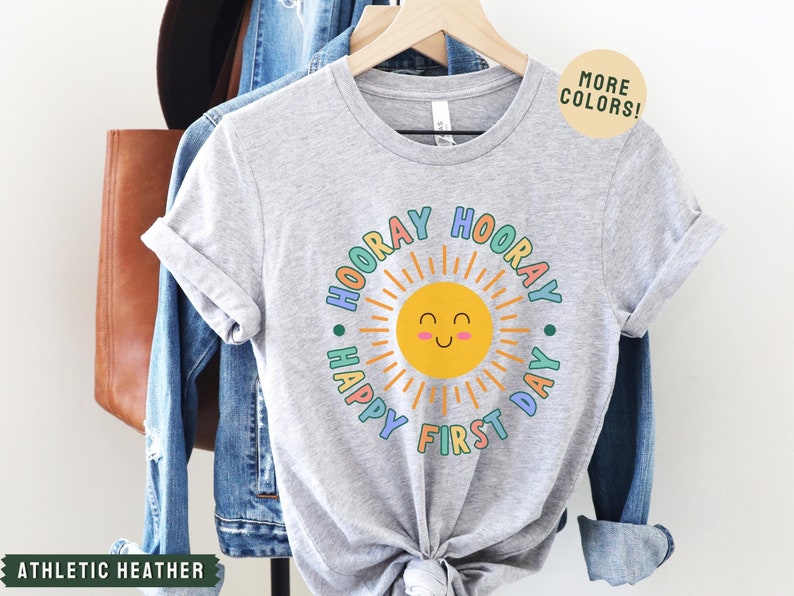K&ouml;nnte beinhalten: Graues T-Shirt mit einem bunten Sonnen-Motiv und dem Text "Hooray Happy First Day" in einem Kreis um die Sonne.