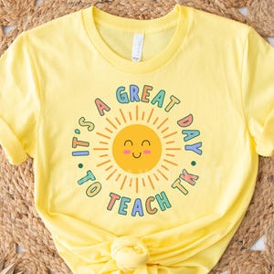 May include: Yellow t-shirt with the text "It's a great day to teach TK" in a circular design. The design includes a smiling sun with colorful accents. The shirt is made of a soft material.
