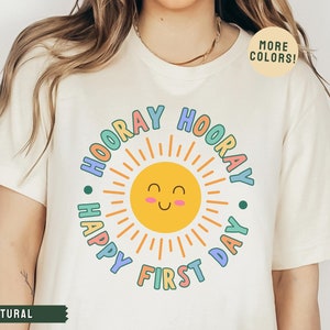 K&ouml;nnte beinhalten: Ein wei&szlig;es T-Shirt mit einer bunten Grafik einer Sonne mit einem l&auml;chelnden Gesicht und dem Text "Hooray Happy First Day".