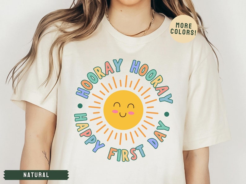 K&ouml;nnte beinhalten: Ein wei&szlig;es T-Shirt mit einer bunten Grafik einer Sonne mit einem l&auml;chelnden Gesicht und dem Text "Hooray Happy First Day".