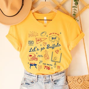 May include: A gold-colored t-shirt with a graphic design featuring the text "Let's go Buffalo" and various football-themed illustrations in red, white, and blue. The shirt includes the number "716" and the word "MAFIA".
