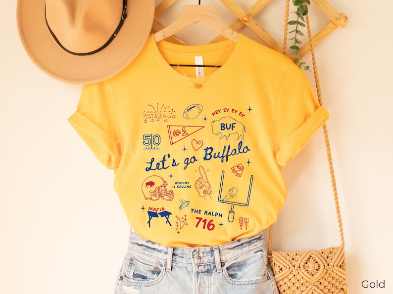 May include: A gold-colored t-shirt with a graphic design featuring the text "Let's go Buffalo" and various football-themed illustrations in red, white, and blue. The shirt includes the number "716" and the word "MAFIA".