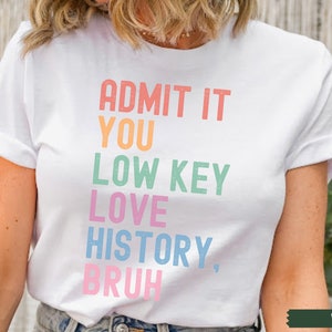 Puede incluir: Camiseta blanca con el texto "ADMIT IT YOU LOW KEY LOVE HISTORY, BRUH" en colores arco&iacute;ris.