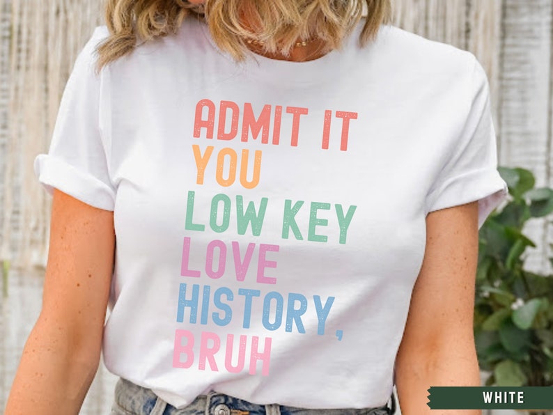 Puede incluir: Camiseta blanca con el texto "ADMIT IT YOU LOW KEY LOVE HISTORY, BRUH" en colores arco&iacute;ris.