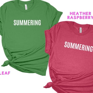 May include: Two t-shirts with the word "SUMMERING" printed in white on the front. The shirt on the left is green and the shirt on the right is a heather raspberry color.