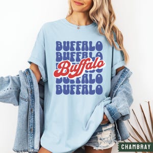 May include: A light blue t-shirt with a retro-style graphic that says "Buffalo" in red and blue letters. The word "Buffalo" is repeated in a stacked pattern on the shirt.