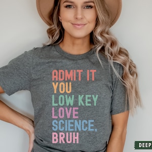 May include: A dark heather grey t-shirt with the text "ADMIT IT YOU LOW KEY LOVE SCIENCE, BRUH" in rainbow colors.