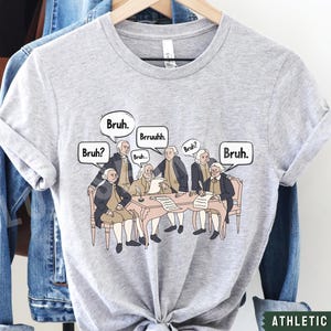 Peut inclure: Un t-shirt gris avec un graphique des P&egrave;res fondateurs assis autour d'une table avec des bulles de dialogue qui disent "Bruh." Le texte "Athletic Heather" est imprim&eacute; en bas du t-shirt.