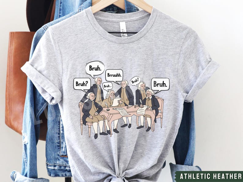 Peut inclure: Un t-shirt gris avec un graphique des P&egrave;res fondateurs assis autour d'une table avec des bulles de dialogue qui disent "Bruh." Le texte "Athletic Heather" est imprim&eacute; en bas du t-shirt.