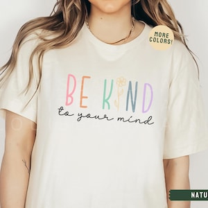 May include: A white t-shirt with the text "BE KIND to your mind" in colorful letters with a flower graphic. The text "MORE COLORS!" is in a green circle on the shirt.