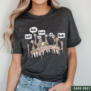 Peut inclure: T-shirt gris fonc&eacute; chin&eacute; avec un motif repr&eacute;sentant les P&egrave;res fondateurs assis autour d'une table avec des bulles de dialogue qui disent "Bruh." et "Bruh?"