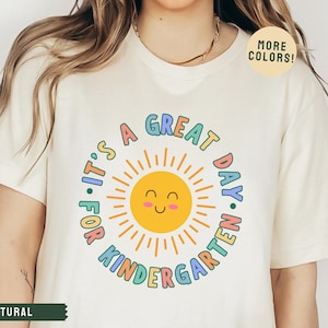 May include: A white t-shirt with a colorful graphic that says "It's a Great Day for Kindergarten" with a smiling sun in the center.
