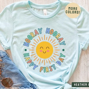 K&ouml;nnte beinhalten: Hellblaues T-Shirt mit einer bunten Grafik einer l&auml;chelnden Sonne und dem Text "Hooray Happy First Day".