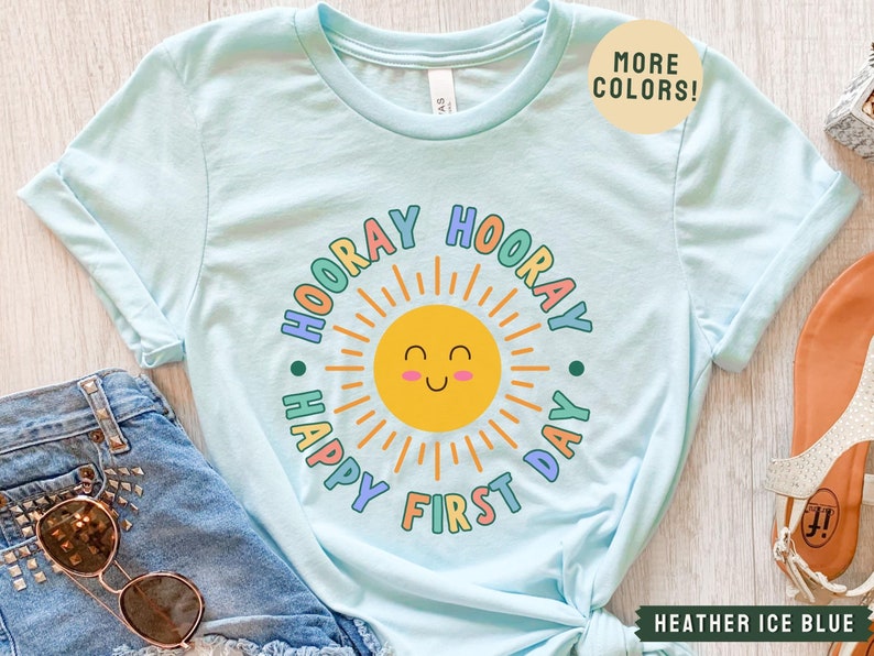 K&ouml;nnte beinhalten: Hellblaues T-Shirt mit einer bunten Grafik einer l&auml;chelnden Sonne und dem Text "Hooray Happy First Day".
