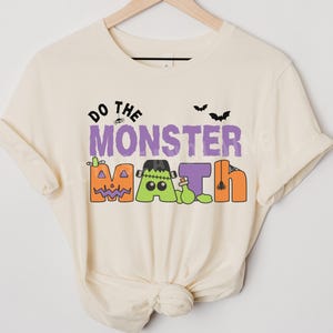 May include: Cream-colored t-shirt with the phrase "Do the Monster Math" in a playful, colorful design. The design features Halloween-themed elements like pumpkins, Frankenstein's monster, and bats, perfect for the spooky season.