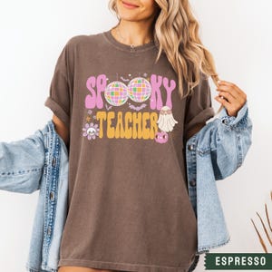 Peut inclure: Un t-shirt marron avec le texte "Spooky Teacher" en lettres roses et jaunes. Le design comprend des boules disco, des chauves-souris, un fant&ocirc;me et un cr&acirc;ne.
