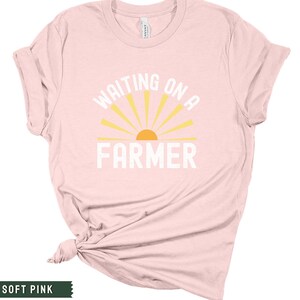 Może przedstawiać: R&oacute;żowy t-shirt z napisem "WAITING ON A FARMER" w kolorze białym i ż&oacute;łtym graficznym słońcem.