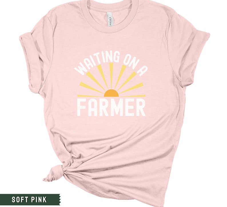 Może przedstawiać: R&oacute;żowy t-shirt z napisem "WAITING ON A FARMER" w kolorze białym i ż&oacute;łtym graficznym słońcem.