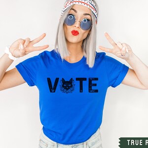 Puede incluir: Una camiseta azul real con un gr&aacute;fico de gato negro y la palabra "VOTE" en letras negras.