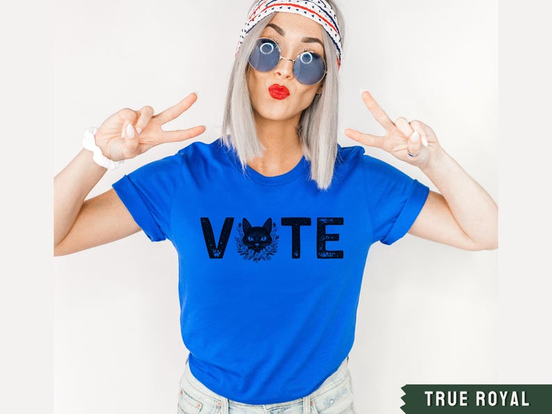 Puede incluir: Una camiseta azul real con un gr&aacute;fico de gato negro y la palabra "VOTE" en letras negras.