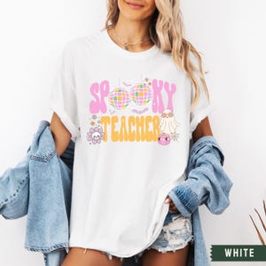 Peut inclure: T-shirt blanc avec un motif rose et jaune qui dit "Spooky Teacher" avec une boule disco, un fant&ocirc;me et des fleurs.