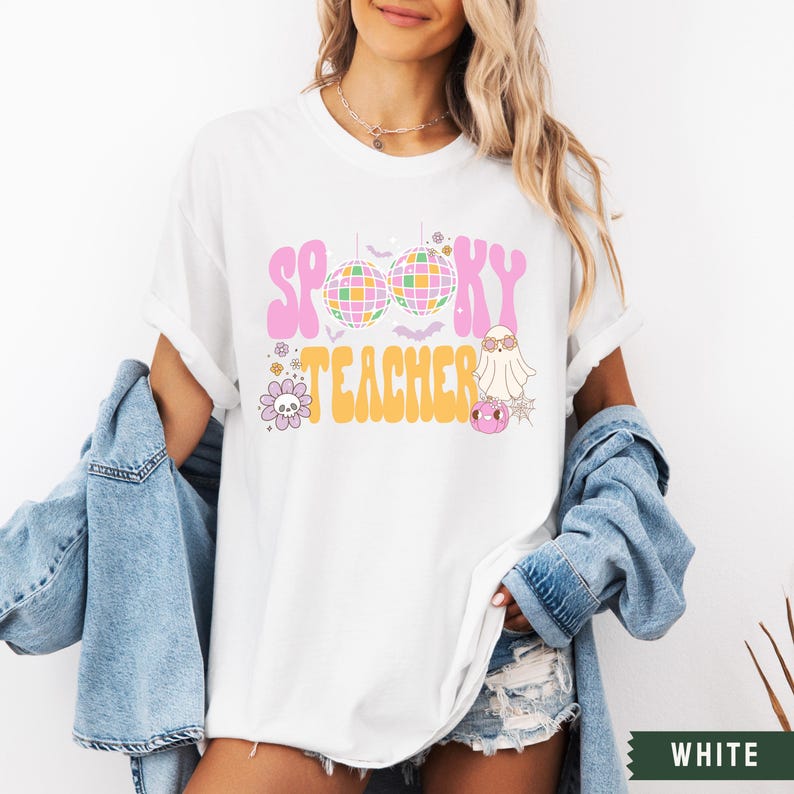 Peut inclure: T-shirt blanc avec un motif rose et jaune qui dit "Spooky Teacher" avec une boule disco, un fant&ocirc;me et des fleurs.