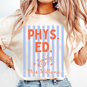 May include: Ivory-colored t-shirt with a graphic that says "PHYS. ED." in orange letters on a blue and white striped background. Below the text is a whistle graphic and the name "Miss Williams".
