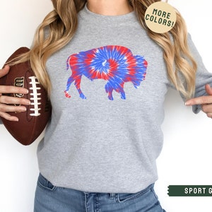 May include: Sport grey sweatshirt featuring a tie-dye buffalo design in red, white, and blue. The sweatshirt has long sleeves and a crew neck. The image also includes a football and text that says "MORE COLORS!" and "SPORT GREY".