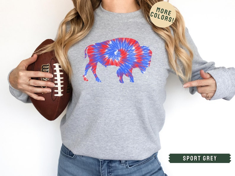 May include: Sport grey sweatshirt featuring a tie-dye buffalo design in red, white, and blue. The sweatshirt has long sleeves and a crew neck. The image also includes a football and text that says "MORE COLORS!" and "SPORT GREY".