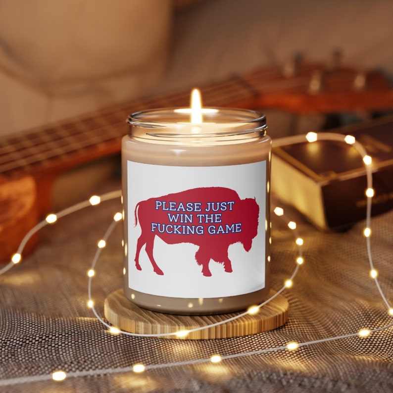 May include: A brown candle with a white label that says "PLEASE JUST WIN THE FUCKING GAME". The label also features a red silhouette of a buffalo. The candle is lit and sitting on a wooden coaster.