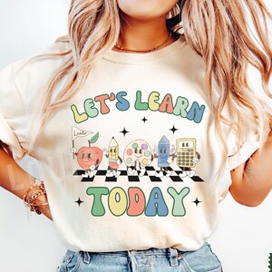 May include: A white t-shirt with the text "Let's Learn Today" in colorful, retro-style lettering. The design features cartoon characters of a pencil, apple, paint palette, ruler, and calculator.