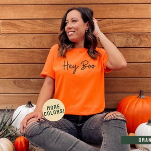 May include: A woman wearing a bright orange t-shirt with the text "Hey Boo" in black script. She is sitting on the ground with pumpkins around her. The t-shirt is available in other colors.