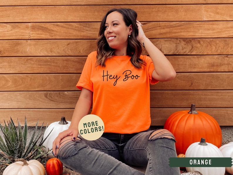 May include: A woman wearing a bright orange t-shirt with the text "Hey Boo" in black script. She is sitting on the ground with pumpkins around her. The t-shirt is available in other colors.