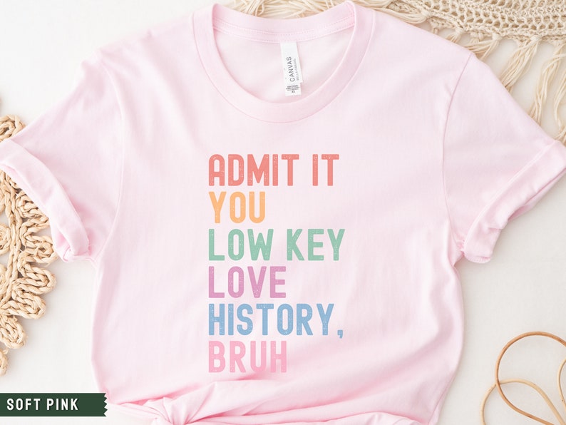 Puede incluir: Una camiseta rosa suave con el texto "ADMIT IT YOU LOW KEY LOVE HISTORY, BRUH" impreso en colores arco&iacute;ris.