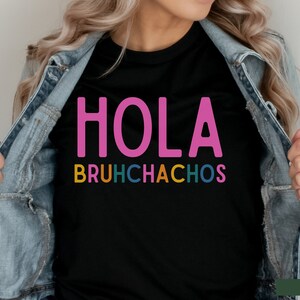 May include: Black t-shirt with the text "HOLA BRUHCHACHOS" in pink, yellow, green, blue, and orange letters.