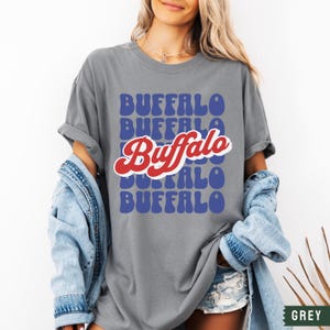 May include: A grey t-shirt with a retro-style graphic design featuring the word "Buffalo" repeated in blue and red letters.