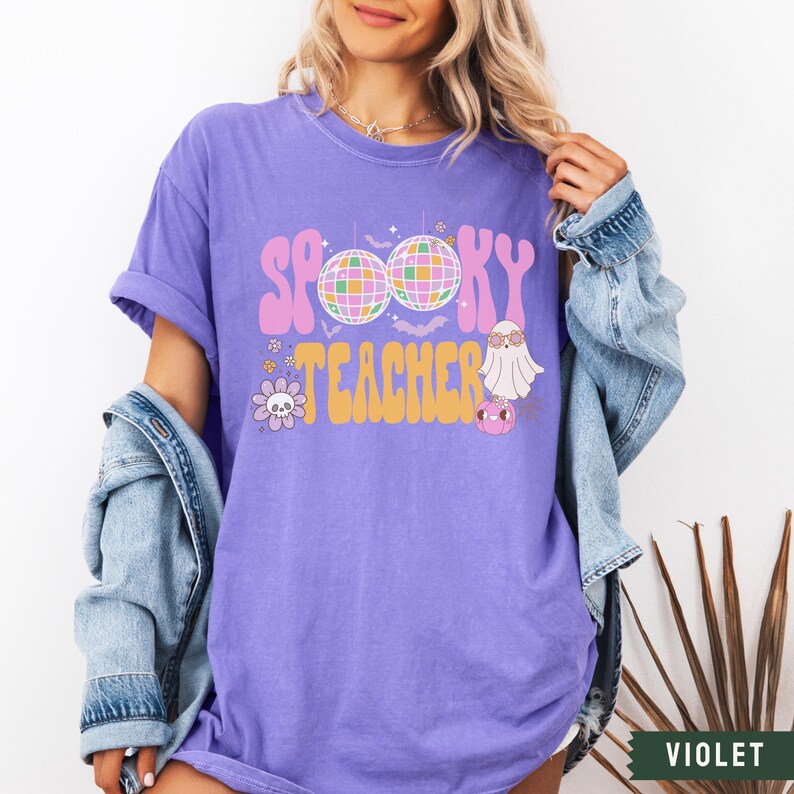 Peut inclure: T-shirt violet avec le texte "Spooky Teacher" dans une police r&eacute;tro. Le design comprend un fant&ocirc;me, des boules disco et d'autres &eacute;l&eacute;ments sur le th&egrave;me d'Halloween.