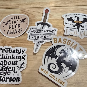 Puede incluir: Una colección de pegatinas con varios diseños y texto. Una pegatina dice "WELL THE FUCK AWARE" con siluetas de dragones. Otra presenta una espada y las palabras "VIOLENT LITTLE THING". Otras pegatinas incluyen gráficos de dragones y frases.