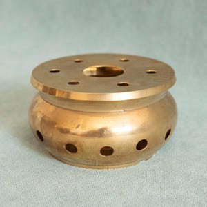 May include: A golden-colored incense burner with a round base and a lid. The lid has a central hole and several smaller holes around the edge. The body of the burner has several circular openings. The object is made of metal.