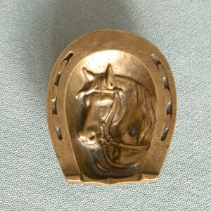 May include: A brass-colored metal object shaped like a horseshoe with a raised relief of a horse head in the center. The horse is depicted wearing a bridle, and the horseshoe frame features several nail holes along its curved outer edges.