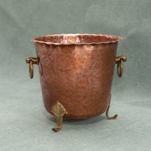 May include: A hammered copper bucket with a scalloped rim, featuring two brass ring handles on the sides and three decorative brass feet shaped like shells. The metal surface displays a warm, reddish-brown tone with a textured, dimpled finish.