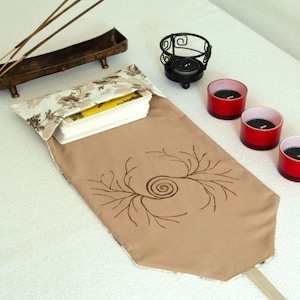 May include: A tan fabric tarot card pouch featuring a brown spiral branch design, with a floral patterned flap holding a deck of cards. Nearby are three red votive candle holders, a black metal candle holder, and a long bronze incense burner.